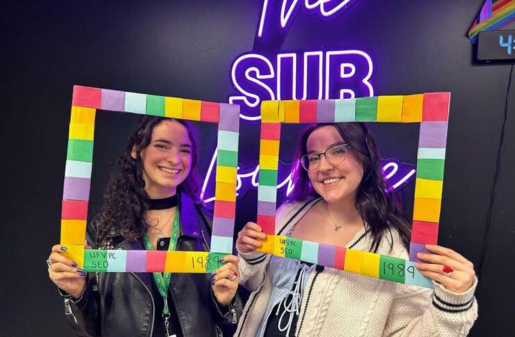 Two ladies holding up a multicoloured frame in front of the SUB lounge sign