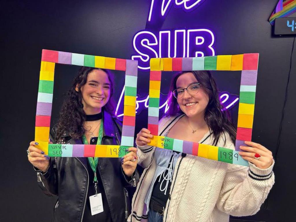 Two ladies holding up a multicoloured frame in front of the SUB lounge sign