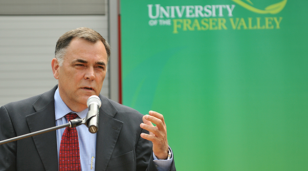 Dr. Darryl Plecas speaking into a mircrophone that is mounted on a podium outside with a university of the fraser valley banner in the background