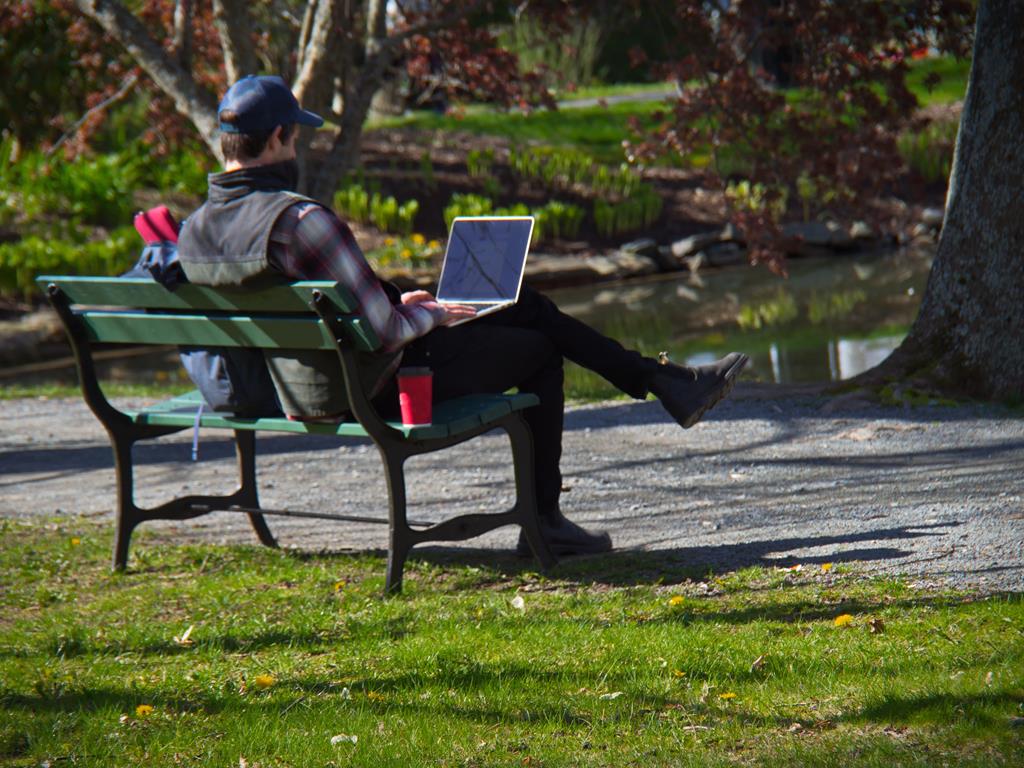 A man with a hat sitting on a bench with a laptop on his lap