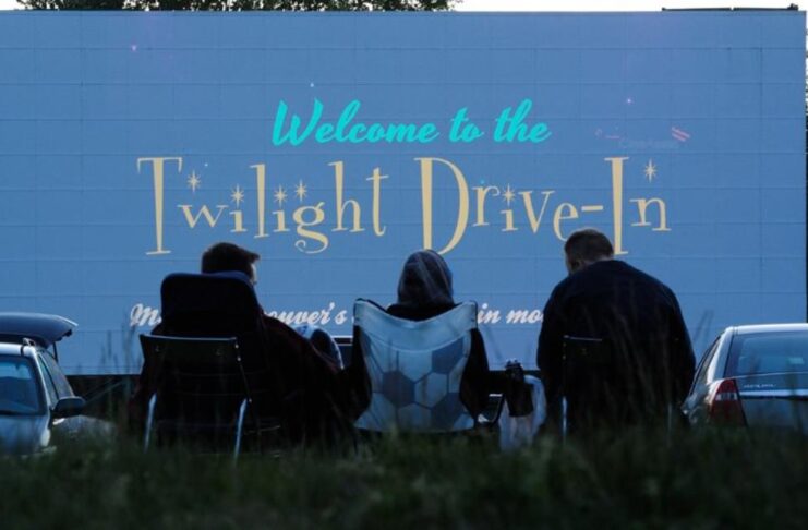 Three people sitting in front of a movie screen outside. The screen says "Welcome to the Twilight Drive In"
